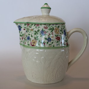 May include: A white ceramic creamer with a floral pattern and a green lid. The creamer has a raised design on the bottom half and a handle.