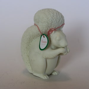 May include: White porcelain squirrel figurine with a pink ribbon around its head. The squirrel is holding a nut in its paws. A green tag with the text "Easter 2021" is attached to the ribbon.