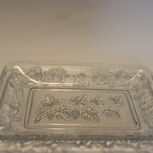 May include: A clear glass rectangular serving tray with a grape and leaf design. The tray has a beaded edge and a raised grape vine pattern in the center and around the border.