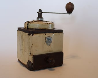 May include: A vintage white and brown manual coffee grinder with a wooden handle and a metal crank. The grinder has a metal drawer and a label with a lion logo that says "Remarkable".
