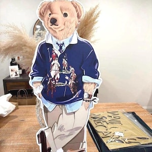 May include: A cardboard cutout of a bear wearing a navy blue polo shirt with a polo player design, a light blue collared shirt, and khaki pants. The bear has a watch on its wrist and is wearing brown shoes. The cutout is standing on a wooden table.