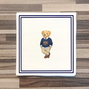 May include: A square white paper napkin featuring a double navy blue border and a central illustration of a tan teddy bear wearing a navy blue sweater with a polo player graphic, a light blue collared shirt, tan trousers, and brown shoes.