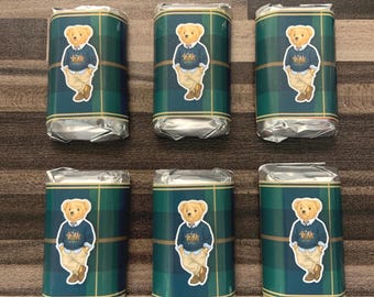 Op de afbeelding: Zes rechthoekige chocoladerepen verpakt in zilverfolie en bedekt met een groen en blauw geruit papieren etiket. Elk etiket is voorzien van een gecentreerde illustratie van een lichtbruine teddybeer in een marineblauwe trui en lichte broek.