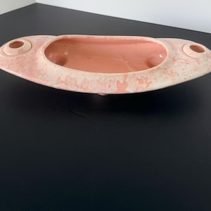 May include: A vintage, oval-shaped ceramic planter in a soft pink hue with a speckled, off-white exterior. The planter features two circular openings on either end and a central, elongated basin. The interior is a solid pink color.