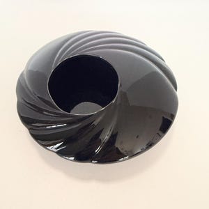 May include: A glossy black ceramic bowl featuring a distinct spiral or swirl pattern radiating from the center opening. The object has a smooth, reflective surface with curved, concave ridges that create a circular, shell-like appearance.
