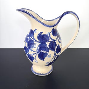 May include: Cream-colored ceramic pitcher with a blue floral design. The pitcher has a rounded body, a curved handle, and a spout. The blue floral pattern includes leaves and vines. The rim and base are trimmed with blue.
