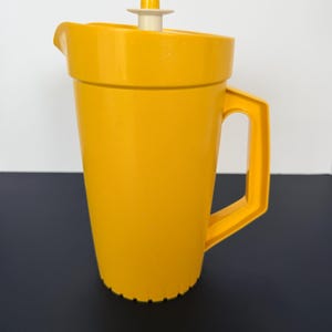 May include: A bright yellow plastic pitcher with a handle and a lid. The pitcher has a wide, cylindrical body and a spout for pouring. The lid is white and yellow. The pitcher is likely used for serving beverages.
