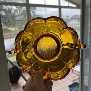 May include: A vintage amber-colored glass dish with a floral petal design. The dish has a central, textured circle and two small handles. The edges of the dish have a detailed embossed pattern. The dish is held up against a window.