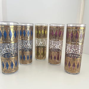 May include: A set of five tall cylindrical drinking glasses featuring gold filigree patterns. Each glass has a band of diamond shapes in blue, yellow, or purple, alternating with a central section of intricate gold floral scrollwork designs.