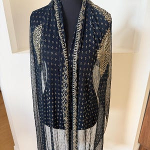 May include: A sheer black shawl with gold geometric patterns and trim. The shawl is draped over a black mannequin, showcasing its flowing design. The gold accents create a striking contrast against the dark fabric, highlighting the intricate details.