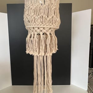 May include: A beige macrame pendant light fixture. The light has a cylindrical shape with intricate knotted patterns and long fringe. The fixture is suspended from a ceiling mount, showcasing a bohemian style.
