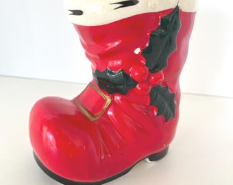 May include: A ceramic Santa boot-shaped vase, primarily red with a white cuff and black accents. The boot features a gold buckle detail and a sprig of holly with red berries. The vase is designed for decorative use.