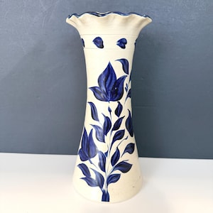 May include: A tall, off-white ceramic vase with a flared, ruffled rim and a blue floral design. The vase features hand-painted blue flowers and leaves, creating a decorative and artistic appeal. The vase is suitable for displaying flowers or as a decorative accent.