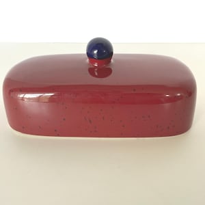 May include: A rectangular ceramic butter dish with a glossy red finish featuring small black speckles throughout. The lid has a smooth, rounded top and is topped with a small, spherical dark blue knob handle centered on the top surface.