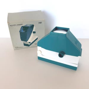 May include: A vintage teal and white Pacer Junior 35mm filmstrip viewer with its original box. The viewer has a teal top, white base, and a lens. The box is light gray with the product name and image printed on it.