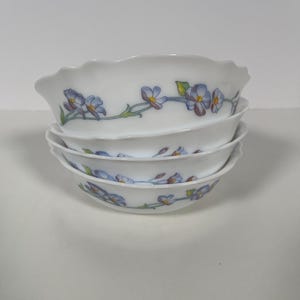 May include: A stack of four white ceramic bowls with a floral design. The bowls have a scalloped edge and feature a delicate pattern of blue and purple flowers with green leaves.