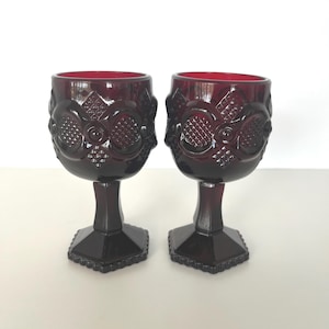 May include: Two ruby red vintage goblets. Each glass features a detailed, embossed pattern around the bowl and a faceted base. The stem is a solid, dark red color. The glasses are displayed on a white surface.