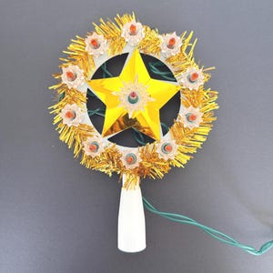May include: A circular tree topper featuring a central gold star surrounded by a ring of ten small, clear, orange-tinted light bulbs. The frame is decorated with gold tinsel and includes a white plastic base for mounting and a green electrical cord.