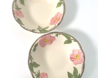 May include: Two round ceramic bowls with a cream-colored base, featuring a hand-painted floral pattern of pink flowers with yellow centers and green leaves. The bowls have a scalloped edge with a thin brown rim, providing a decorative finish.