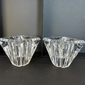 May include: Two clear glass candle holders with a geometric, faceted design. The holders are square shaped and have a wide, open top.
