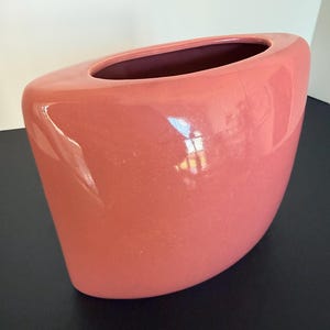 May include: A glossy, coral-pink ceramic vase with a wide, curved, semi-oval silhouette. The vessel features a smooth, reflective surface and a wide, elongated opening at the top, showcasing a deep, hollow interior with a dark-toned inner lining.