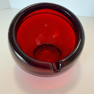 May include: A round, ruby-red glass ashtray with a smooth, glossy finish. The ashtray has a deep bowl shape with a small notch for resting cigarettes. The interior is a vibrant red, and the exterior is a darker shade of red.