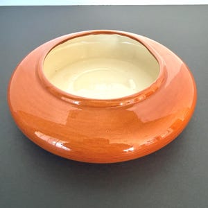 May include: A low-profile, circular ceramic bowl featuring a glossy, vibrant orange exterior finish and a smooth, cream-colored interior. The vessel has a wide, rounded body with a thick, slightly raised rim surrounding the central opening.