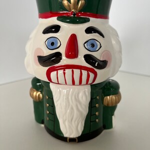 May include: A ceramic nutcracker figurine with a green hat, red and white trim, and a white beard. The nutcracker has a red nose, black mustache, and blue eyes.