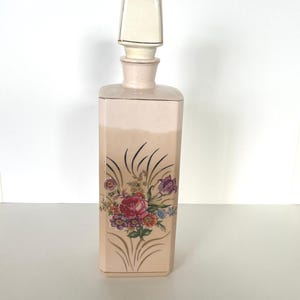 May include: A rectangular ceramic decanter in a soft peach tone featuring a floral bouquet illustration with red, purple, and orange blossoms. The bottle includes a matching square stopper with a thin gold rim, and the base is decorated with gold line accents.