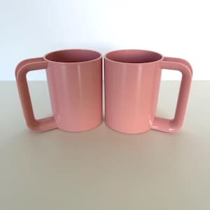 May include: Two matching pink mugs with large, rectangular handles. The mugs are a solid, matte pink color and sit on a light-colored surface. The mugs are empty and ready to be filled with a beverage.