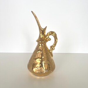 May include: A gold-colored, vintage-style pitcher with a long, curved spout and a decorative handle. The pitcher has a textured surface and a bulbous body. The overall design is ornate and elegant, suitable for decorative purposes.
