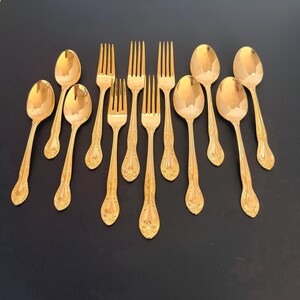 May include: A collection of gold-colored silverware, including spoons and forks, arranged on a dark surface. The silverware has ornate detailing on the handles, suggesting a vintage or antique style. The spoons and forks are arranged in a fan-like pattern.