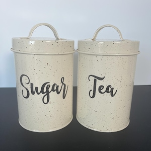 May include: Two matching cream-colored metal canisters with speckled detailing. One canister reads "Sugar" in a black script, and the other reads "Tea." Each has a curved handle on top.