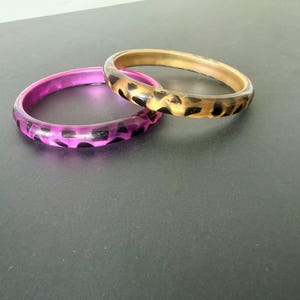 May include: Two bangle bracelets. One is a translucent magenta with black spots, and the other is a gold-toned color with black spots. The bracelets are made of a hard, shiny material and are circular in shape.