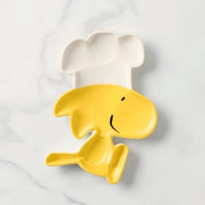 Può includere: Un piatto in ceramica giallo a forma di personaggio dei cartoni animati, con un cappello da chef bianco. Il piatto ha una faccia sorridente ed è progettato per servire cibo. Il piatto è di colore giallo brillante.