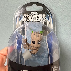 Puede incluir: Una figura de NECA Scalers Potted Groot en un paquete de plástico transparente. La figura es un pequeño personaje marrón con forma de árbol, con hojas verdes y una maceta gris. El embalaje incluye el texto "SCALERS" y "AGES 8+".