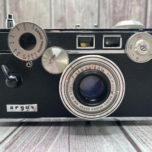May include: A vintage black Argus camera with silver dials and a lens. The camera has the text "RANGE FINDER" and "argus" on the front. The lens reads "f/3.5 30 MM ARGUS COATED CINTAR". The camera is on a wooden surface.