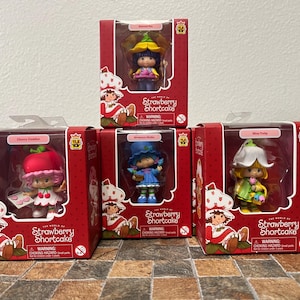 May include: Five Strawberry Shortcake character figurines in their original red boxes. Each box features a different character, including Cherry Cuddler and Miss Huckleberry Pie. The boxes have the text "Strawberry Shortcake" and a warning about choking hazards.