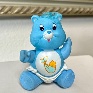 May include: A light blue Care Bear figurine with a white diaper. The bear has a smiling face, black eyes, and a tuft of blue hair. The bear's belly features a graphic of a baby in a stork's embrace with a star.