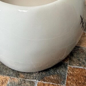 May include: A white ceramic bowl with a rounded form. The bowl has a smooth surface with a few visible lines and a stitched design on the side. The bowl is on a tiled surface.