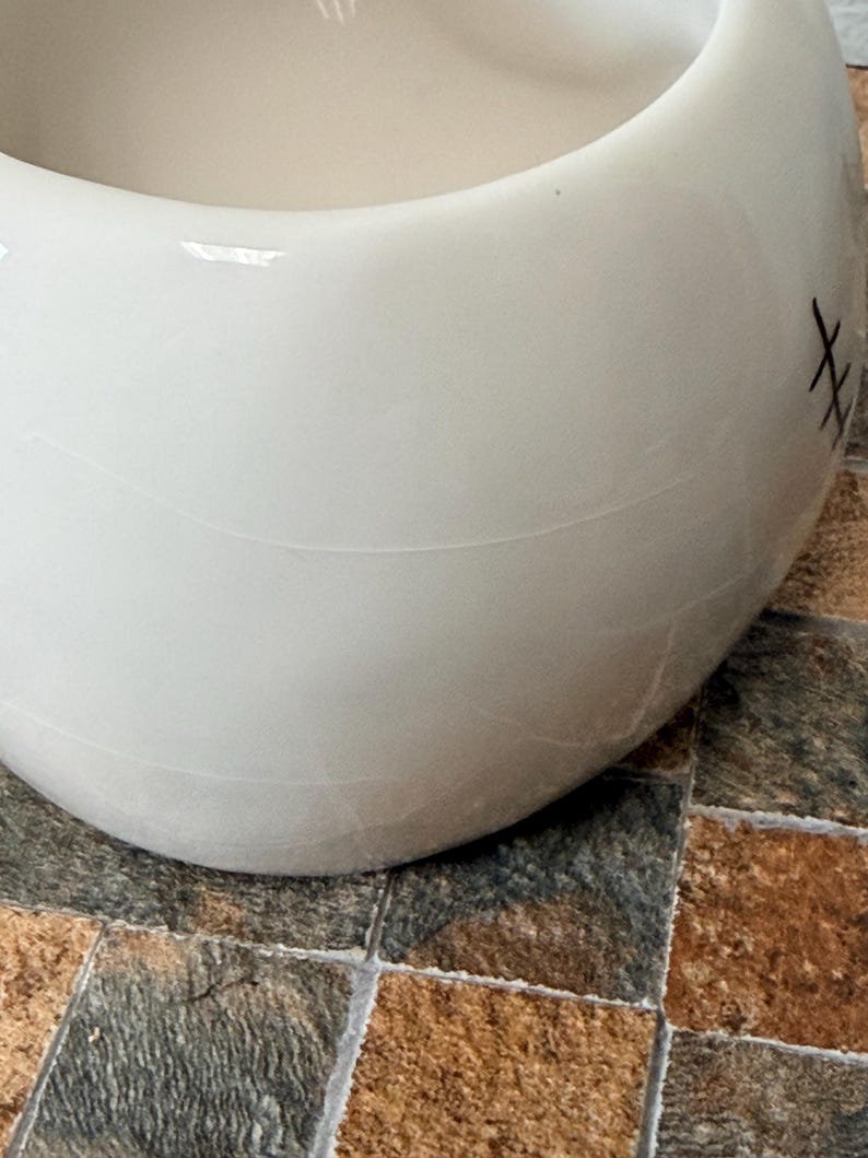 May include: A white ceramic bowl with a rounded form. The bowl has a smooth surface with a few visible lines and a stitched design on the side. The bowl is on a tiled surface.