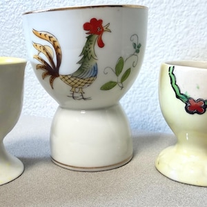 May include: Three ceramic egg cups of varying designs. The left cup is off-white with yellow interior accents. The center cup is white, featuring a colorful rooster illustration and green vines. The right cup is pale yellow with a green and red floral trim.