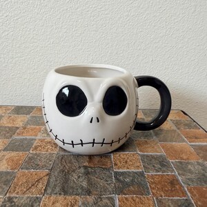May include: White ceramic mug featuring Jack Skellington's face from The Nightmare Before Christmas. The mug has large black eye sockets, a small nose, and a stitched smile. The handle is black.