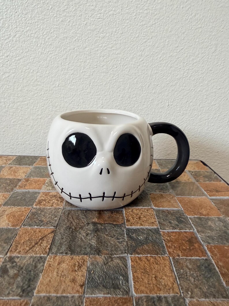 May include: White ceramic mug featuring Jack Skellington's face from The Nightmare Before Christmas. The mug has large black eye sockets, a small nose, and a stitched smile. The handle is black.