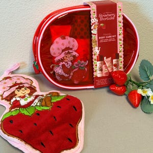 Puede incluir: Un kit de cuidado corporal de viaje Strawberry Shortcake en un estuche rojo translúcido con cremallera y una bolsa de felpa con forma de fresa a juego que muestra al personaje. El kit incluye loción, aceite, crema de manos y gel de baño.