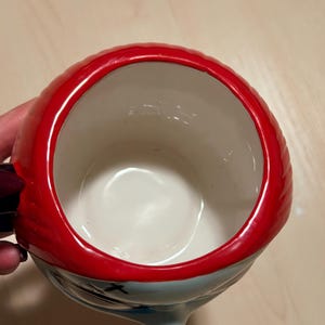 May include: A ceramic mug with a character design. It has red hair, a light blue face, and black accents. The mug has a white interior and a red rim. The handle is black and purple. The mug is designed for drinks.