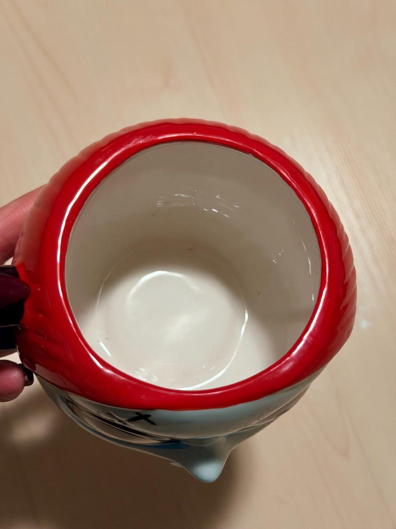 May include: A ceramic mug with a character design. It has red hair, a light blue face, and black accents. The mug has a white interior and a red rim. The handle is black and purple. The mug is designed for drinks.