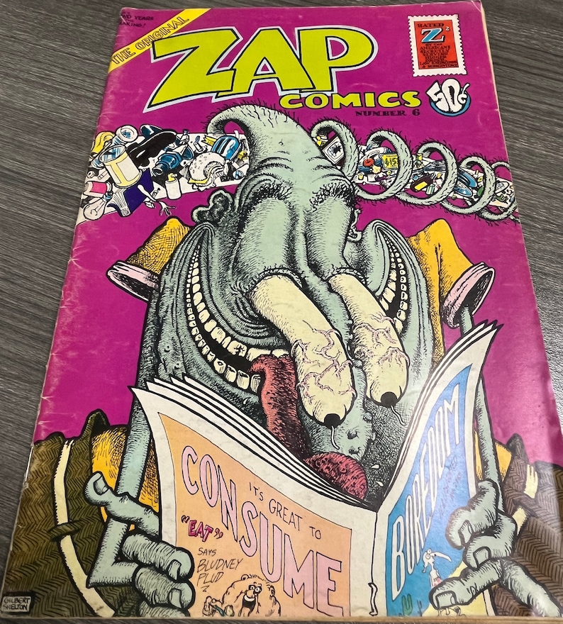 May include: A vintage comic book cover featuring a green monster with a large mouth and a tongue sticking out. The monster is reading a newspaper with the headline "It's Great to Consume" and the text "Eat" in quotation marks. The comic book is titled "Zap Comics" and is number 6. The cover is pink with a yellow border.