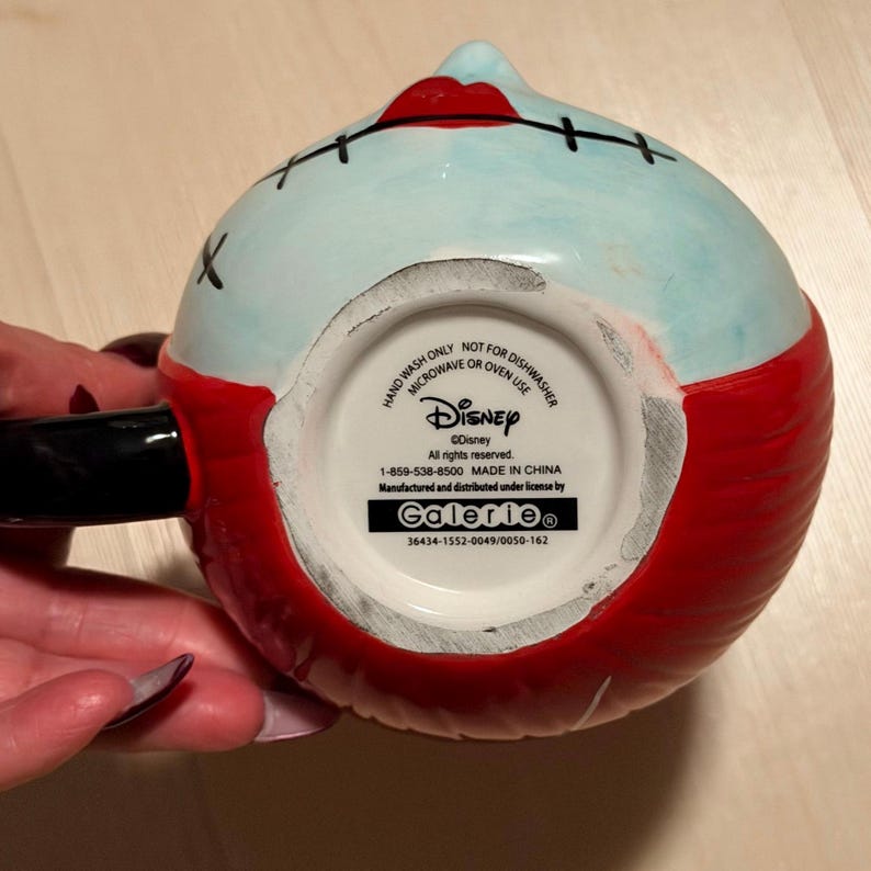 May include: A ceramic mug with a character design, featuring a light blue face, red hair, and stitched details. The mug has a black handle and a red base. The bottom of the mug has text that reads "Disney" and "Galerie".