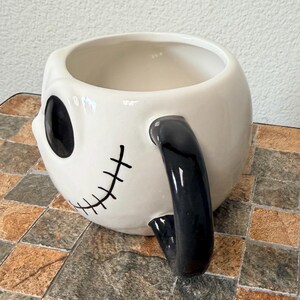 May include: A white ceramic mug with a black handle and skull-like facial features. The mug features a large black eye and a stitched smile. A unique and decorative item.
